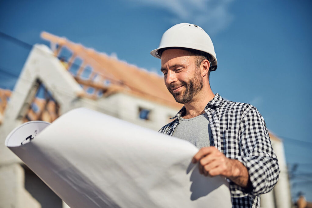 Ein Mann mit Bauhelm sieht zufrieden auf einen Bauplan, im Hintergrund ist ein Haus, dass sich im Bau befindet